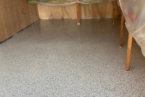 Light gray flake floor in a garage with plywood walls, with plastic sheeting draped over the wooden shelves.