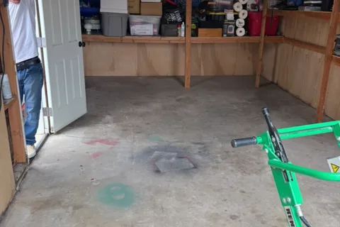 Bare concrete floor with paint stains in a garage with plywood walls and shelves, a green floor machine in front and a person at the open door.