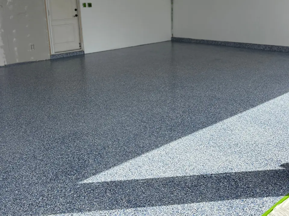 Blue, gray and white flake garage floor with sunlight across it, white walls and a door at the back left.