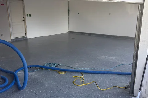 Two-car garage with a gray floor, a blue vacuum hose and yellow extension cords across it, and a door in the white back wall.