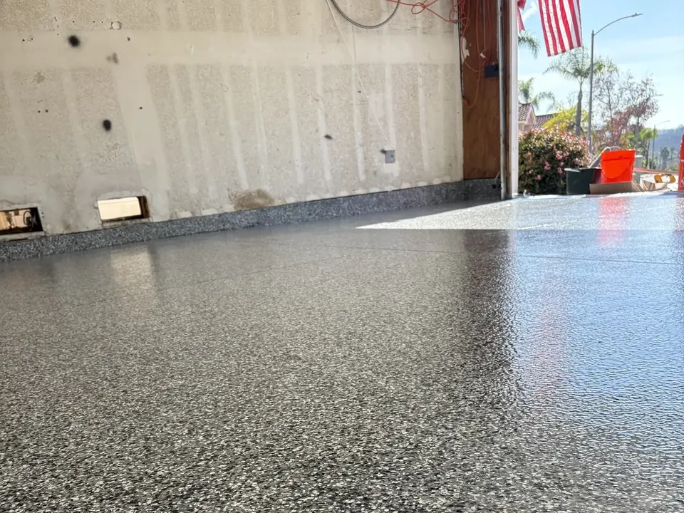 Low-angle view across a glossy black, gray and white flake floor to an open garage door, with an American flag and palm trees outside.