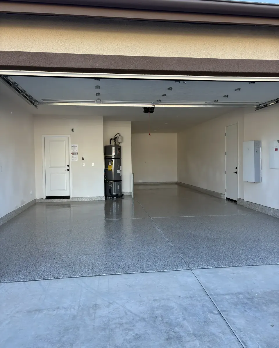 Open two-car garage seen from the driveway, with a gray flake floor, coated stem walls, and a white door and water heater at the back.
