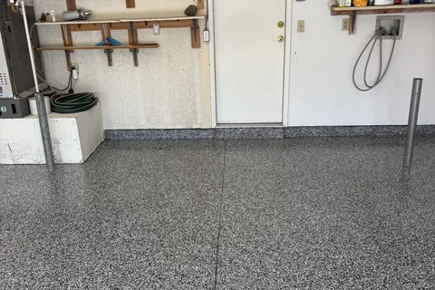 Black, gray and white flake garage floor with straight joint lines, in front of a white door, wooden wall shelves and a furnace.