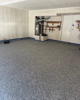 Wide view of a gray flake garage floor with blue painter's tape still along the base of the walls, and a furnace and shelves at the back.