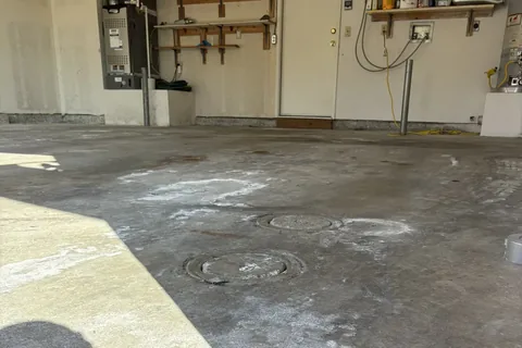 Bare, stained concrete garage floor with two round floor covers and sunlight across the left side; a furnace, shelves and a white door at the back.