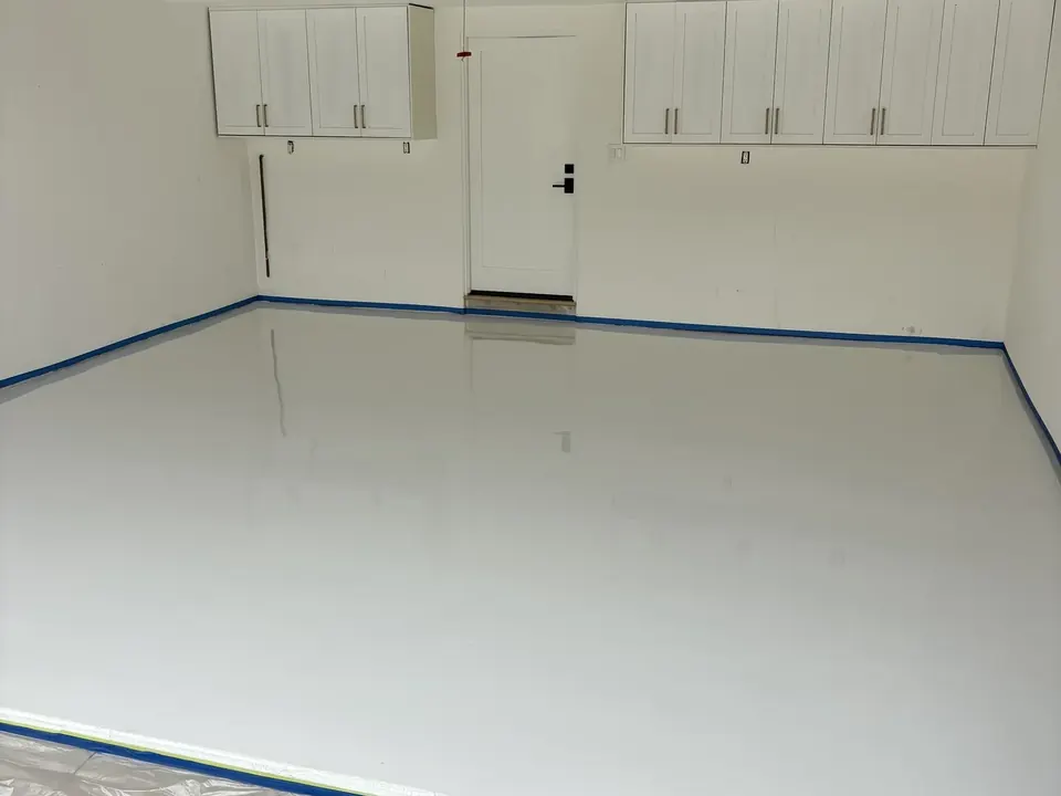 Glossy white garage floor edged with blue painter's tape, white wall cabinets and a white door at the back, and plastic sheeting in front.