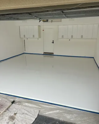 Glossy white garage floor edged with blue painter's tape, white wall cabinets and a white door at the back, and plastic sheeting in front.