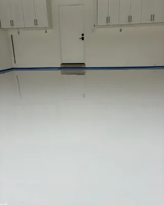 Glossy white garage floor reflecting a white door and white wall cabinets, with blue painter's tape along the base of the wall.