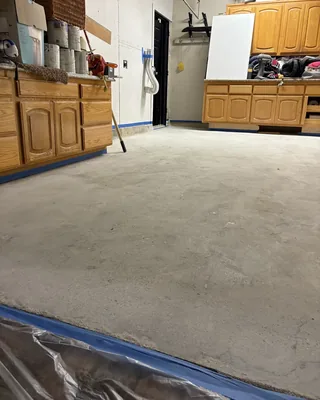 Bare, ground concrete garage floor with the cabinet bases and walls masked in blue tape, and plastic sheeting in front.