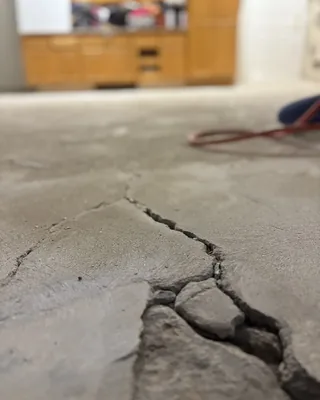 Close-up of a wide crack with loose broken pieces in a bare concrete garage floor, with wooden cabinets blurred behind.