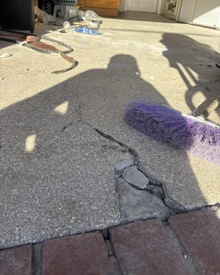 Cracked and broken concrete at a garage entrance beside red brick pavers, with a purple broom and people's shadows on the floor.