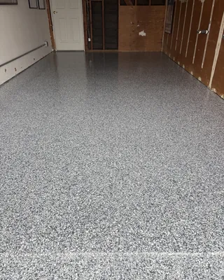 Gray and white flake floor along a narrow garage, with a white door and open wall framing at the back and plywood panels along the right wall.