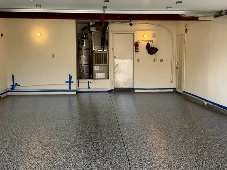 Two-car garage with a gray, black and white flake floor, blue painter's tape along the base of cream walls, and a water heater beside a white door.