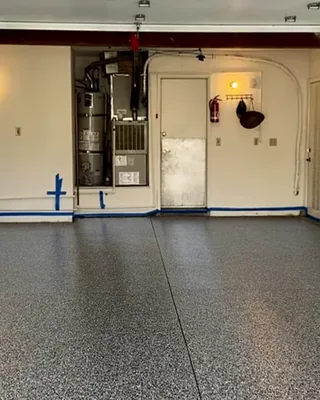 Two-car garage with a gray, black and white flake floor, blue painter's tape along the base of cream walls, and a water heater beside a white door.